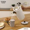 A person is pouring hot water from a white electric kettle into a mug on a wooden countertop, with pastries and a bagel nearby. Produktbild-Vorschau