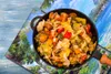 A close-up overhead shot of a pan filled with a colorful vegetable stir-fry, placed on a blue wooden surface with a scenic landscape image underneath. Produktbild-Vorschau