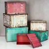 A collection of storage boxes designed to look like industrial shipping containers, stacked and arranged on a white surface against a gray concrete wall. Produktbild-Vorschau