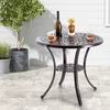 A round metal table with a decorative top and a lower shelf, set on a patio with two glasses of amber liquid, a bottle of wine, and a small vase with a yellow flower. A potted plant is visible to the left. Náhled obrázku produktu