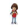 A stylized figurine of a young boy wearing a red vest over a blue plaid shirt, light blue pants, and black and white sneakers, with a green backpack. Podgląd zdjęcia produktu