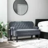 A dark gray velvet bench with a tufted backrest is placed in a bedroom setting next to a bed. A large round mirror hangs on the wall behind the bench, and a potted plant is visible to the left. Náhled obrázku produktu