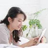 A young woman is lying on a bed, smiling and looking at a tablet while wearing earphones. Produktbild-Vorschau