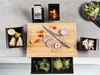 A kitchen counter with a bamboo cutting board, knife, and various ingredients for cooking, including onions, tomatoes, ginger, lemons, mushrooms, broccoli, and sprouts. A smartphone is also visible, likely displaying a recipe. Produktbild-Vorschau