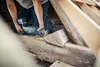 A person is using a reciprocating saw to cut through a wooden beam in what appears to be an attic or old building. Voir les images du produit