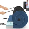 A rolled-up camping mat with a blue fuzzy exterior and a black ribbed interior, shown with straps being pulled by hands. A person is shown in the background wearing the mat in a carrying bag. Náhľad obrázku produktu