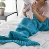 A young girl sits on a bed, wrapped in a teal mermaid tail blanket and holding a stuffed toy. Produktbild-Vorschau