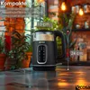 A black electric kettle is shown on a reflective surface with a blurred background of plants and lights. Produktbild-Vorschau