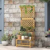 A wooden plant stand with multiple tiers and a trellis, adorned with various potted plants, is placed against a brick wall next to a door. A watering can sits on the ground nearby. Produktbild-Vorschau