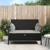 A black poly rattan 2-seater garden bench with gray cushions is placed on a paved patio in front of a wooden fence, with plants and trees in the background. Náhľad obrázku produktu
