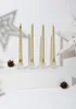 Four gold spiral candles in clear holders on a white surface, with pinecones and star decorations in the background. Anteprima dell’immagine prodotto