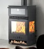 A black wood-burning stove with a glass door and a compartment for logs at the bottom, set against a textured gray wall. Produktbild-Vorschau