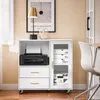 A white sideboard with a printer and drawers on the left, and a glass-doored cabinet on the right, is placed in a room with a lamp and a framed picture on the wall. Produktbild-Vorschau