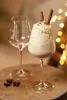 A festive scene with a glass of eggnog and an empty wine glass, decorated with cinnamon sticks and star anise, with bokeh lights in the background. Produktbild-Vorschau