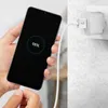 A hand is holding a smartphone that is charging, with the charger plugged into a wall socket. Náhľad obrázku produktu