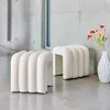 Two white fabric-covered ottomans are placed on a concrete floor in front of a concrete wall with windows. A white vase with orange flowers is to the right of the ottomans. Produktbild-Vorschau