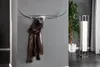 A silver bull skull-shaped coat rack with a brown jacket hanging on it, mounted on a gray wall. A tall potted plant and a floor lamp are visible in the background. Produktbild-Vorschau