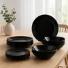 A set of black plates and bowls are neatly stacked on a wooden table, with a vase of eucalyptus in the background. Náhled obrázku produktu