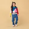 A young girl wearing a colorful sweater and jeans, with a backpack on, stands against a plain yellow background. Produktbild-Vorschau
