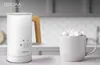 A white milk frother with a wooden handle sits on a countertop next to a mug filled with marshmallows, with a blurred kitchen background. Produktbild-Vorschau