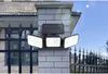 A solar-powered LED light fixture mounted on a brick pillar, with a fence and building in the background. Podgląd zdjęcia produktu