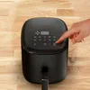 A hand is shown pressing the start/stop button on the digital display of a black air fryer, which is placed on a wooden surface. Produktbild-Vorschau