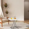 A white folding table with a teapot and cup of tea on it, placed on a rug in a living room next to a chair. The background is a plain wall with a door on the right. Produktbild-Vorschau