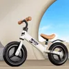 A white balance bike for children is shown against a backdrop of a curved beige structure and a blue sky. Náhled obrázku produktu