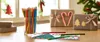 A collection of colored pencils and Christmas-themed gift wrapping supplies on a wooden table. Anteprima dell’immagine prodotto