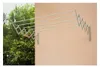 A retractable clothesline mounted on a wall, with trees and foliage in the background on the left side. Náhled obrázku produktu