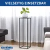 A black metal plant stand with a potted plant on top, placed in a living room setting with sofas and curtains. Produktbild-Vorschau