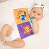 A baby lying on a white surface, holding and looking at a soft fabric book with colorful illustrations. Anteprima dell’immagine prodotto