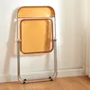 A transparent orange folding chair with a silver metal frame stands against a white wall. Produktbild-Vorschau