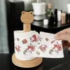 A hand is holding a roll of toilet paper with a Christmas design, featuring Santa Claus and gift boxes. The toilet paper is on a wooden stand with a frog-shaped topper. Náhľad obrázku produktu