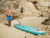 A man is pumping up an inflatable stand-up paddleboard on a sandy beach with rocky cliffs in the background. Produktbild-Vorschau