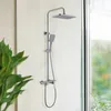 A modern shower fixture with a rain showerhead and handheld sprayer is mounted on a wall next to a large window with greenery visible outside. A lush green plant is in the foreground. Produktbild-Vorschau