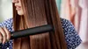 A woman is straightening her long brown hair with a black hair straightener. Náhled obrázku produktu