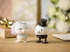 A bride and groom figurine set on a wooden surface with gifts and flowers in the background. Produktbild-Vorschau