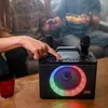 People are gathered around a table, with one person interacting with a karaoke machine that has colorful LED lights. Produktbild-Vorschau