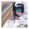 A person wearing white gloves is using a Makita demolition hammer to break concrete, with a pile of rebar in the background. Anteprima dell’immagine prodotto