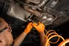 A person wearing orange gloves is using a pneumatic cutting tool on the exhaust system of a car. Produktbild-Vorschau