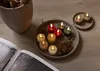 A collection of LED candles arranged on a tray with pinecones, next to an open book and a planter. Produktbild-Vorschau