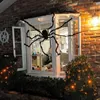 A large, fuzzy black spider decoration is draped across a window and the brick wall of a house, with bushes decorated with orange lights in the foreground. Produktbild-Vorschau