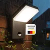 A black LED outdoor wall light with a motion sensor is mounted on a brick wall during a rain shower. A German flag badge with stars is attached to the light. Produktbild-Vorschau