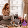 A young woman is sitting by a window, drinking from a mug, with an aroma diffuser emitting mist in the foreground. Voir les images du produit