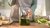A person is using an immersion blender to make pesto in a glass container on a wooden countertop. Anteprima dell’immagine prodotto