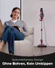 A woman sits on a couch reading a book while a red and silver vacuum cleaner stands upright in the background. Produktbild-Vorschau