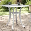 A white square garden table with two cups and saucers on it, placed on a paved patio with greenery in the background. Náhled obrázku produktu