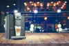 A coffee machine on a wooden table in front of a blurred background of a cafe at night. Produktbild-Vorschau