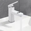 A white kitchen faucet with a digital display is shown in use, with water flowing into a white sink. The background is a gray tiled wall. Produktbild-Vorschau